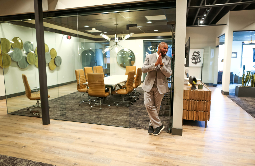 Man in a light suit smiling near a modern glass-walled conference room.