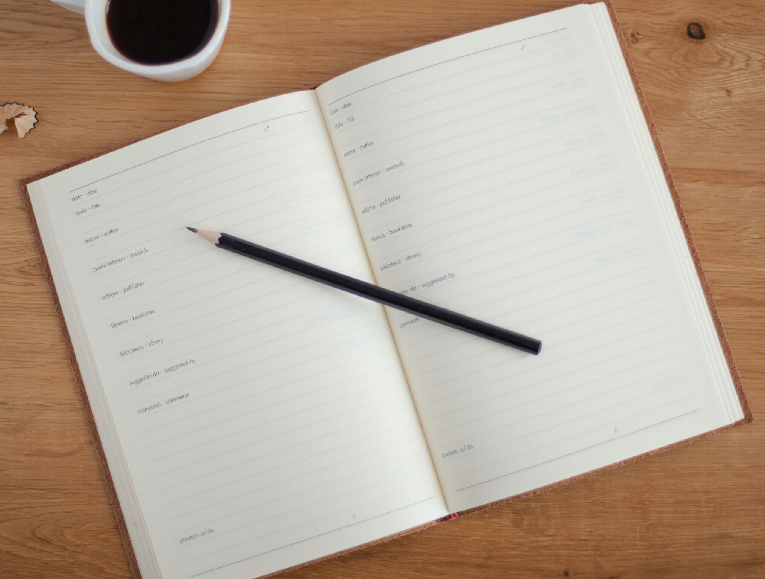 An open notebook with a black pencil and a cup of coffee on a wooden table.